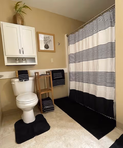 A bathroom with a white toilet, black and white striped shower curtain, black bath mats, and black towels. There is a wooden shelf holding towels next to the toilet, a white cabinet mounted on the wall above the toilet, and a framed botanical print on the wall.