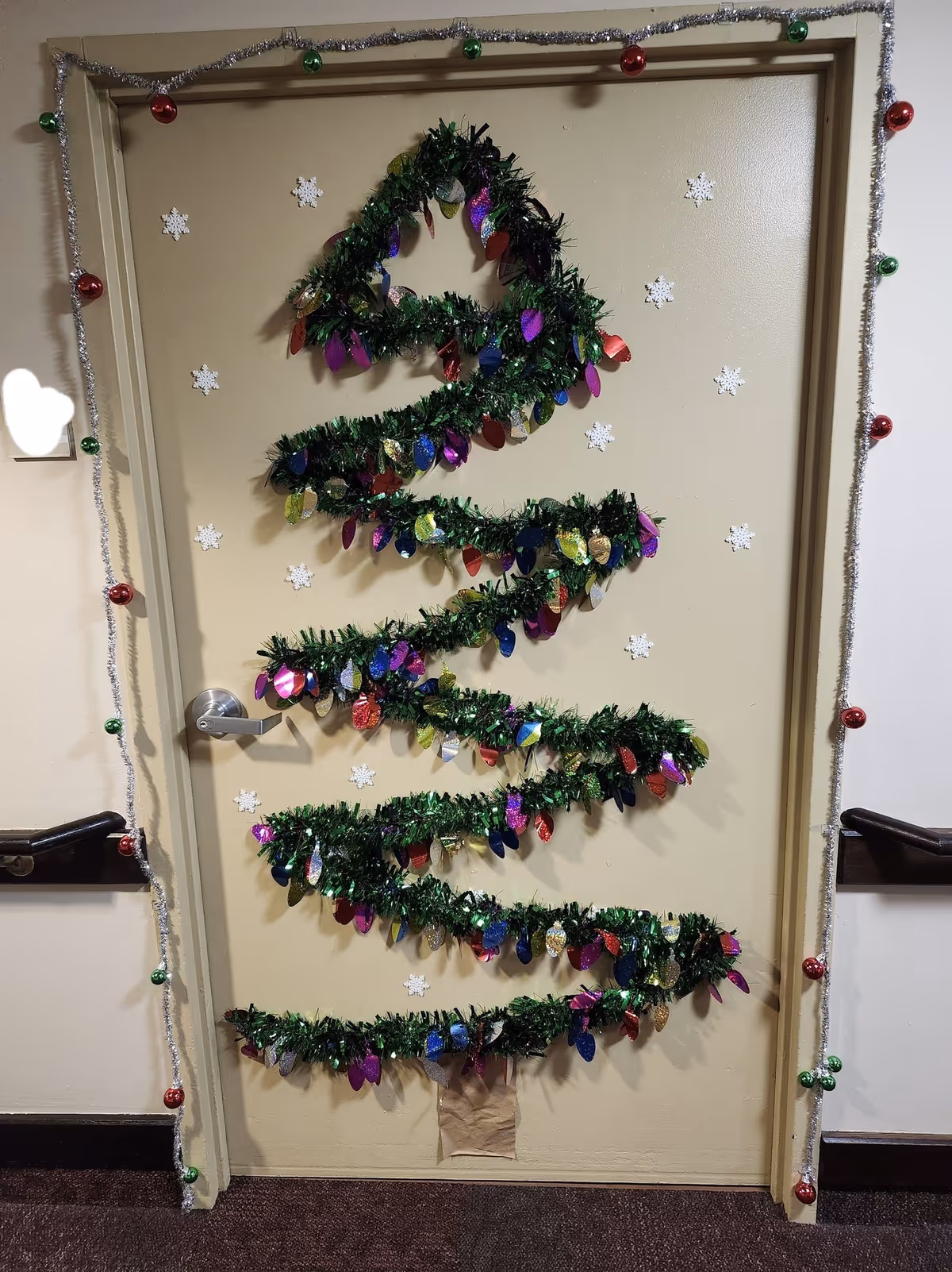 A beige door decorated with a Christmas tree design made from green garland with colorful shiny ornaments attached. The door frame is adorned with silver tinsel garland and red and green baubles. White snowflake decorations are scattered on the door around the tree design.