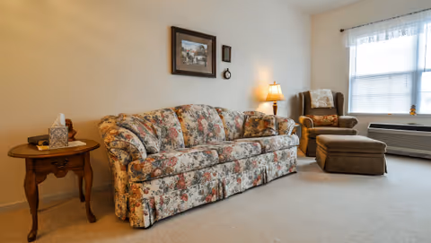 Cozy living room with a floral-patterned sofa, armchair and ottoman, side table with a lamp, and a window.