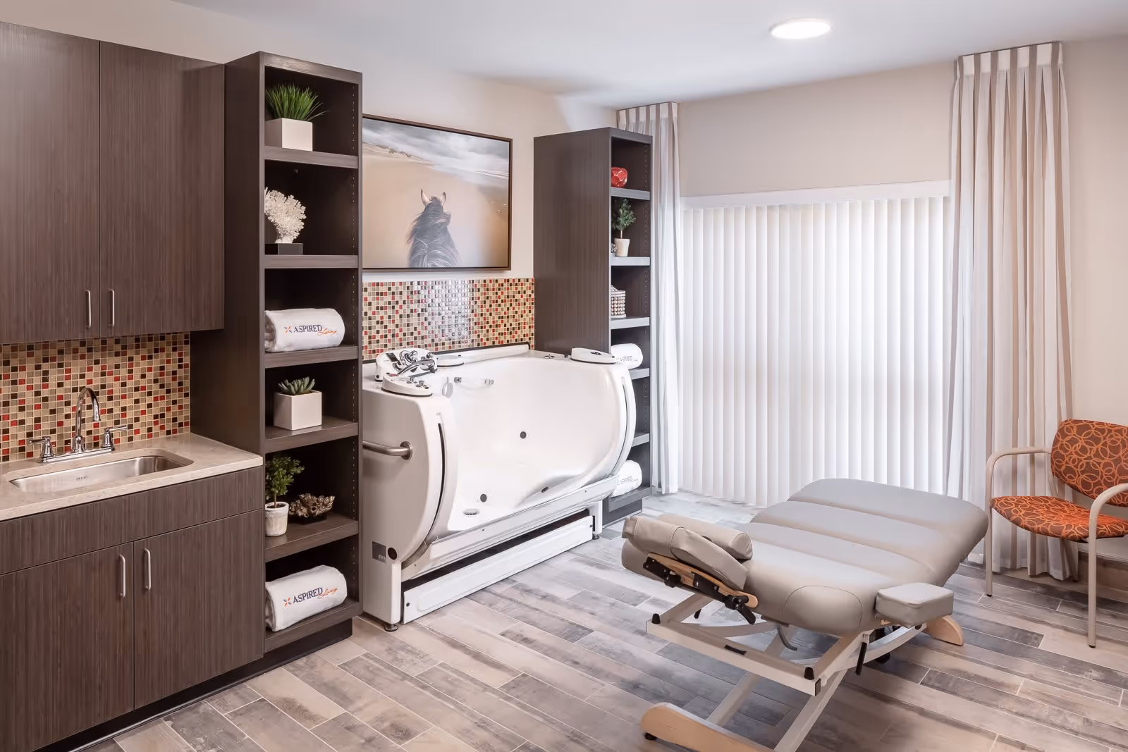 A bright room featuring a white therapeutic bathtub with grab bars, surrounded by dark wood shelving units holding towels and decorative plants. To the left, there is a sink with a mosaic tile backsplash. In front of the tub, there is a padded adjustable therapy table, and to the right, an orange patterned chair sits near a window with vertical blinds and beige curtains.