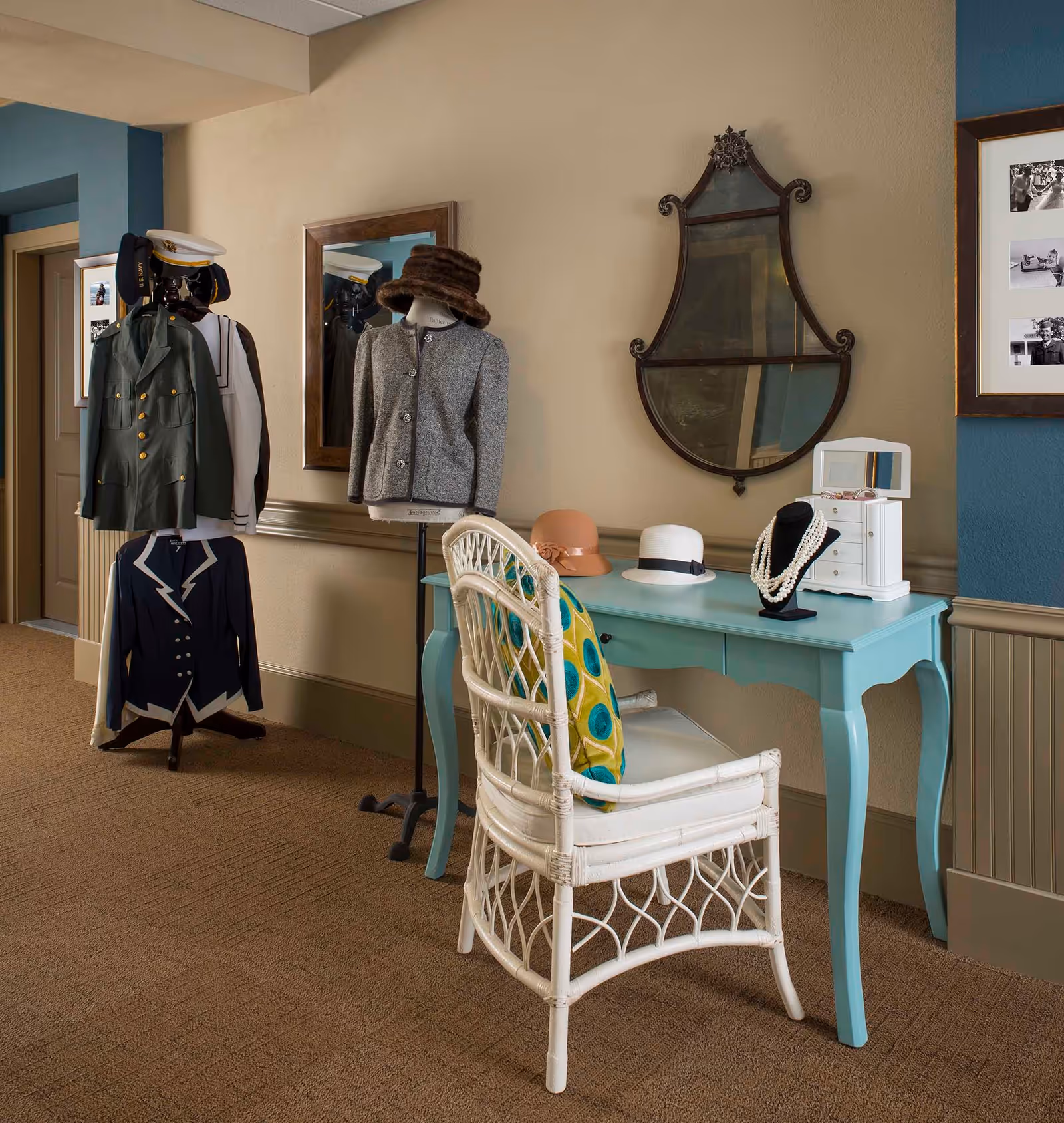 A cozy interior corner featuring a light blue vanity table with a white wicker chair adorned with a colorful cushion. On the table are two hats, a jewelry display with pearl necklaces, and a small white jewelry box with a mirror. Behind the table is an ornate wall mirror. To the left, there are mannequins dressed in vintage clothing and hats, with a framed mirror on the wall reflecting part of the scene.