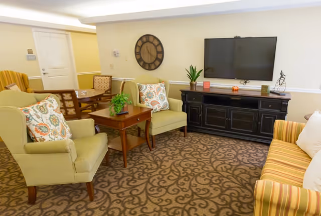 A cozy common living room with upholstered chairs and a striped sofa arranged around a wall-mounted TV and decorative clock.