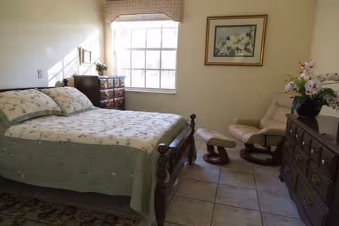 Cozy bedroom with a large bed, dresser, window, and a cushioned chair.