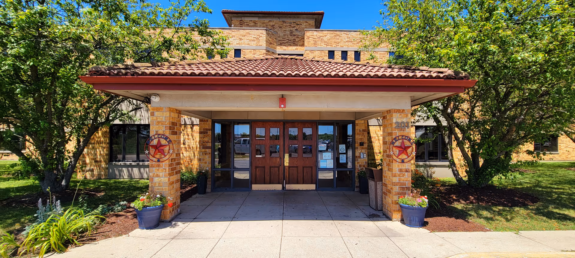 Entrance of a brick building with a covered porch supported by two brick columns. Each column has a decorative star emblem. There are double wooden doors with glass panels and additional glass windows on either side. The area is surrounded by green trees and plants under a clear blue sky.