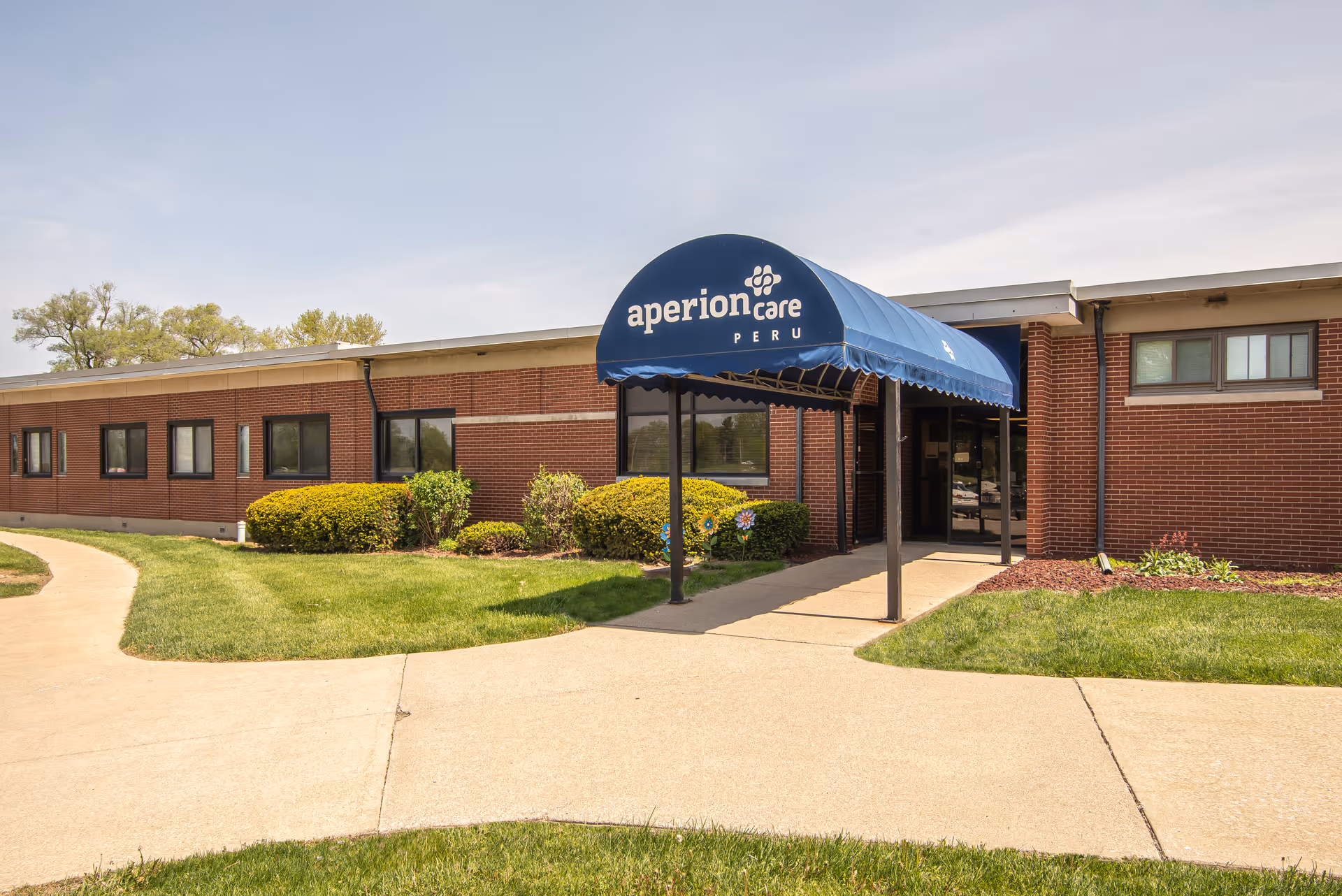 Front entrance of a single-story brick building with a blue awning reading 'aperion care PERU' and a paved walkway.