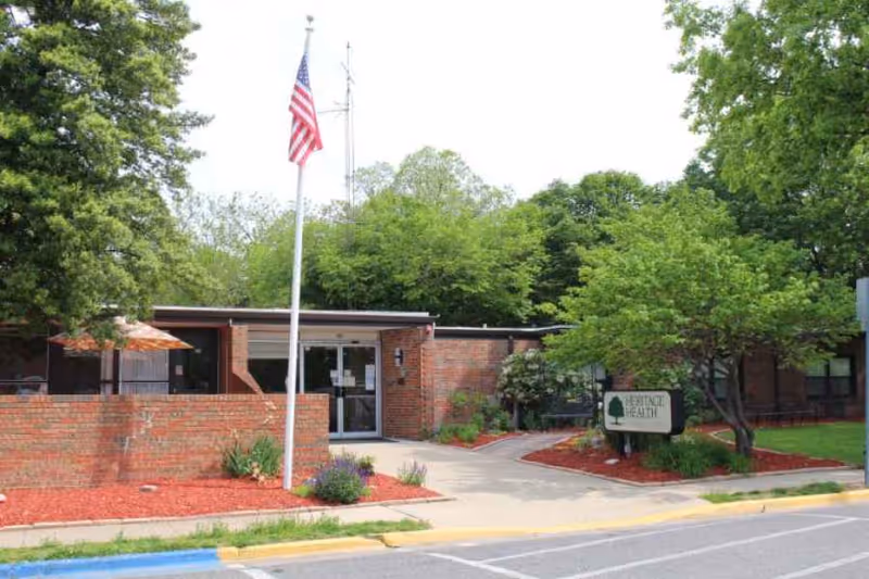 Exterior view of Staunton Health and Rehab Center showing a single-story brick building with a glass entrance door. There is a flagpole with an American flag in front, surrounded by landscaped areas with red mulch, plants, and trees. A sign near the entrance reads 'Advance Health'.