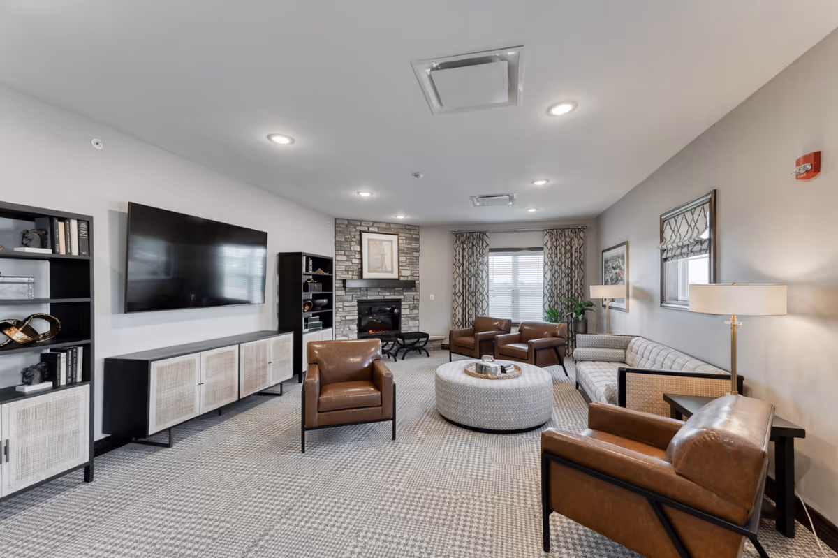 A cozy senior living facility common area with a modern design featuring a large flat-screen TV mounted on the wall, a stone fireplace, several brown leather armchairs, a gray patterned sofa, a round ottoman coffee table, bookshelves, and large windows with patterned curtains allowing natural light.