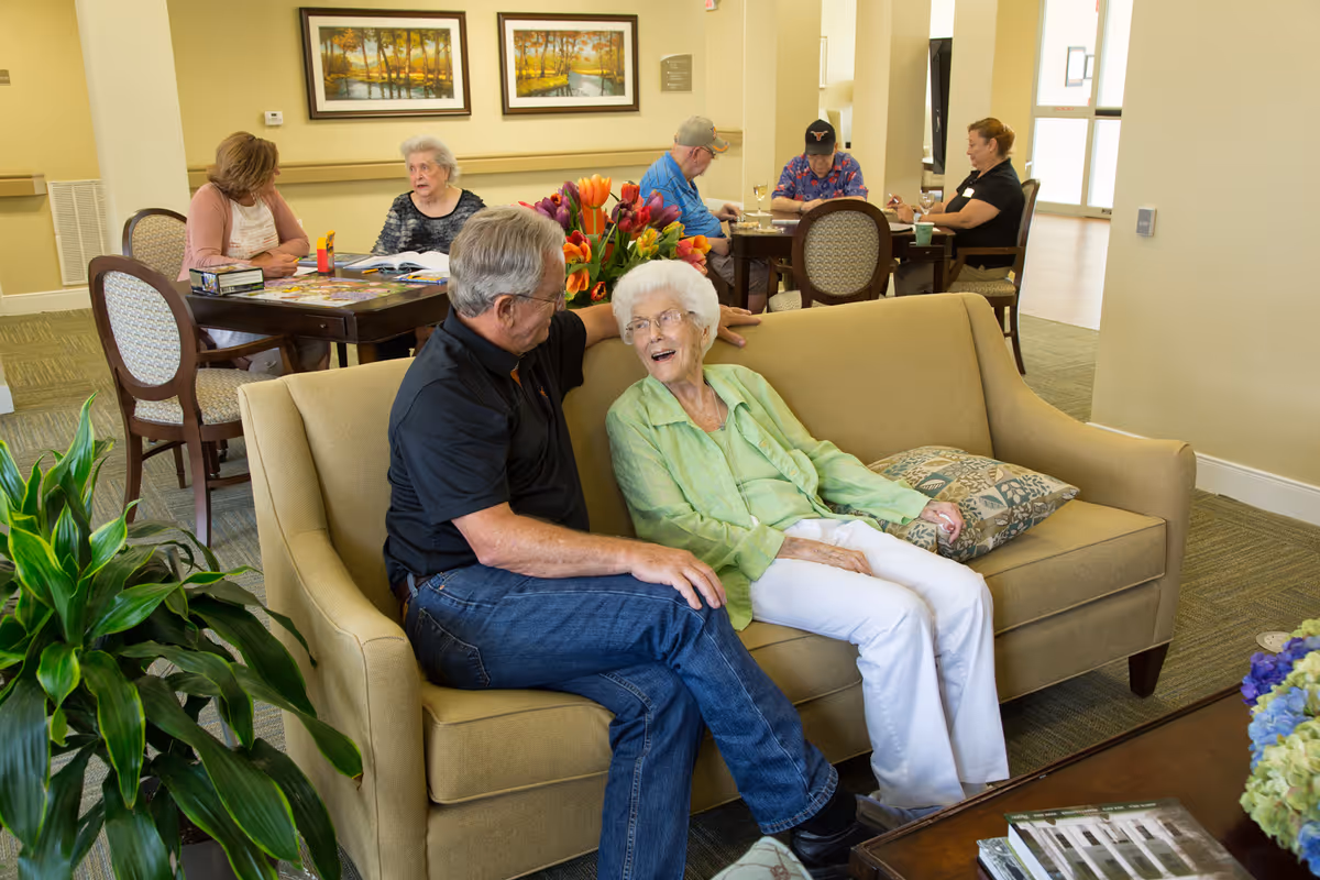 An elderly woman and a man sitting and talking on a beige couch in a common area of a senior living facility. In the background, other elderly people are seated at tables engaged in activities. The room is decorated with plants, flowers, and framed landscape paintings on the wall.