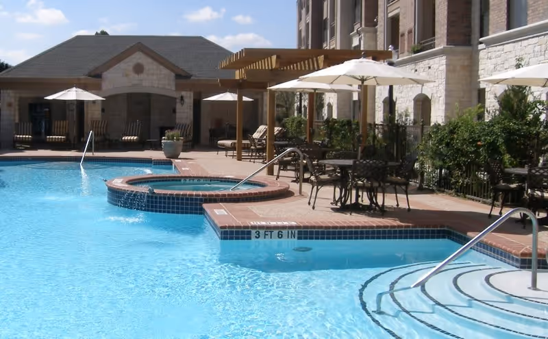 Outdoor swimming pool area with clear blue water, a hot tub, poolside tables with umbrellas, lounge chairs, and a pergola. The pool area is adjacent to a stone building with arched windows and greenery along the fence.