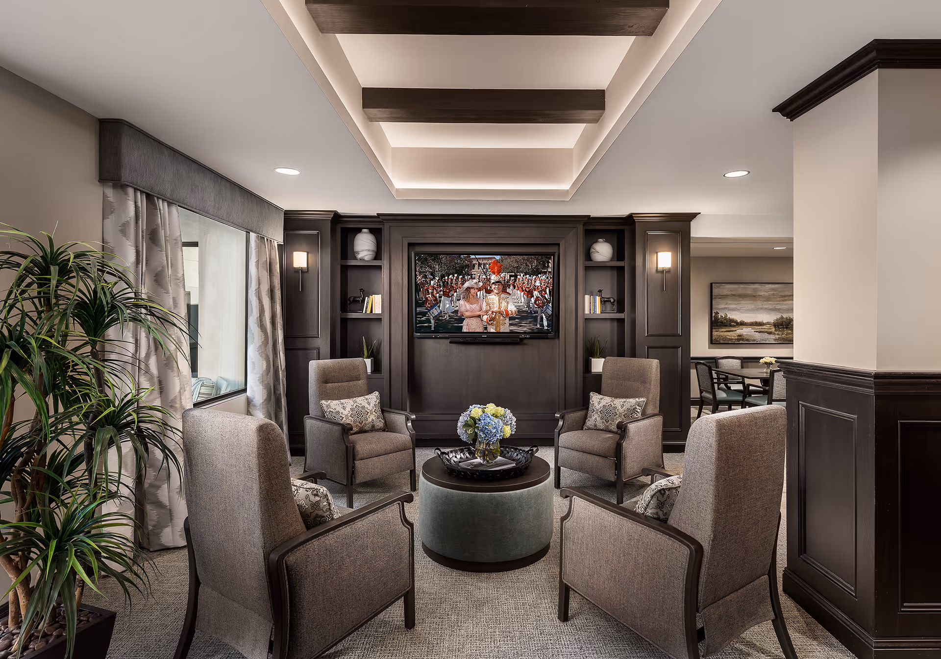 A cozy living room area with four upholstered armchairs arranged around a round ottoman with a flower vase on top. The room features dark wood built-in shelves and cabinetry with a flat-screen TV mounted in the center. There is a large window with curtains on the left side and a potted plant nearby. The ceiling has recessed lighting and decorative beams, and a dining area is visible in the background.