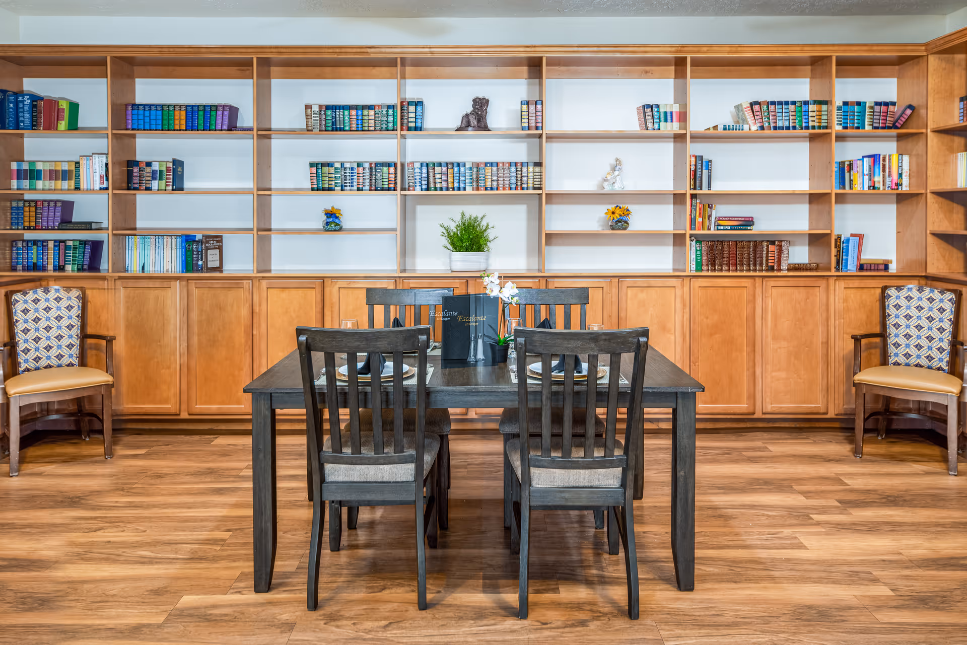 A cozy interior room featuring a wooden dining table with four chairs set with placemats and a small flower centerpiece. Behind the table is a large wooden bookshelf filled with books and decorative items. Two upholstered armchairs with patterned fabric are placed on either side of the bookshelf. The room has wooden flooring and a warm, inviting atmosphere.