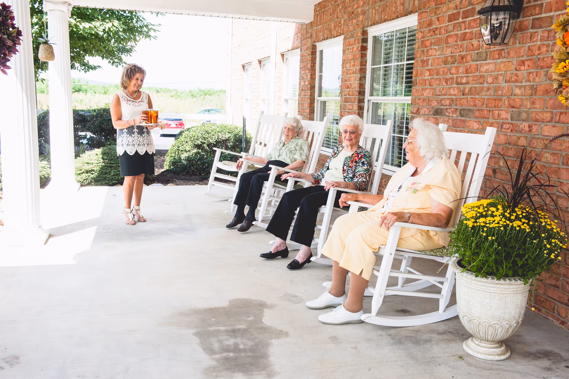 Three elderly women sitting on white rocking chairs on a covered porch with a brick wall behind them. A woman stands nearby holding a tray with drinks. There are potted yellow flowers and greenery around the porch area.