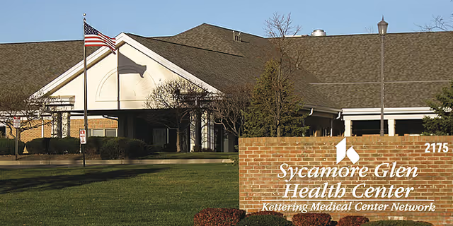 Front exterior of Sycamore Glen Health Center showing the brick entrance sign, American flag, lawn, and building facade.