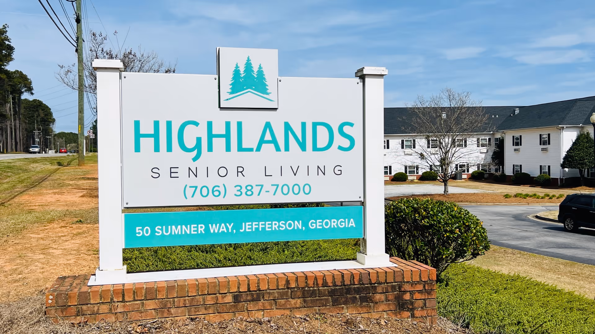 White roadside sign reading 'HIGHLANDS SENIOR LIVING' with phone number and address in front of a two-story senior living building.