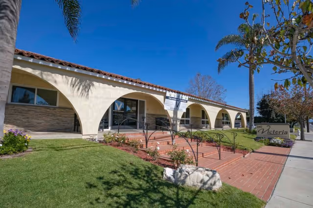 Front entrance of a single-story Mediterranean-style building with arched walkways, tiled roof, landscaped lawn and a sign reading "Victoria".