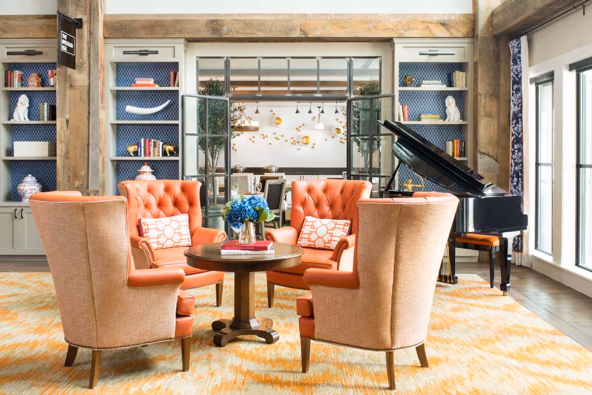 A cozy living room area with four orange tufted armchairs arranged around a round wooden table with a vase of blue flowers and books. Behind the chairs, there are built-in shelves with decorative items and books, and a black grand piano is positioned near large windows letting in natural light. The room features wooden beams and a patterned yellow and white rug.