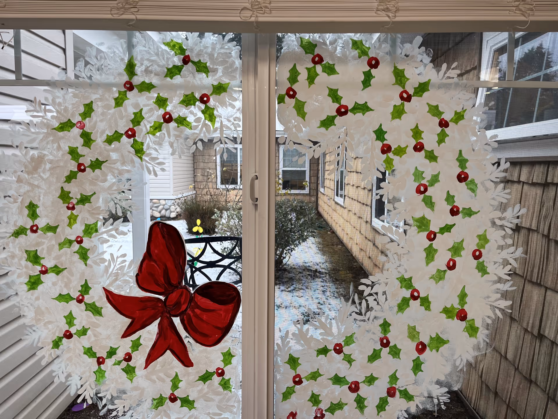 Glass sliding door/window painted with a large white holiday wreath decorated with green holly, red berries and a big red bow, looking out to a small courtyard.