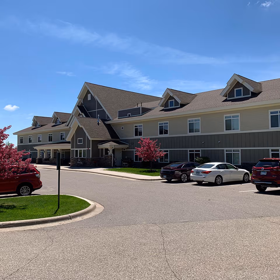 Exterior view of a two-story senior living facility with parked cars and flowering trees under a blue sky.