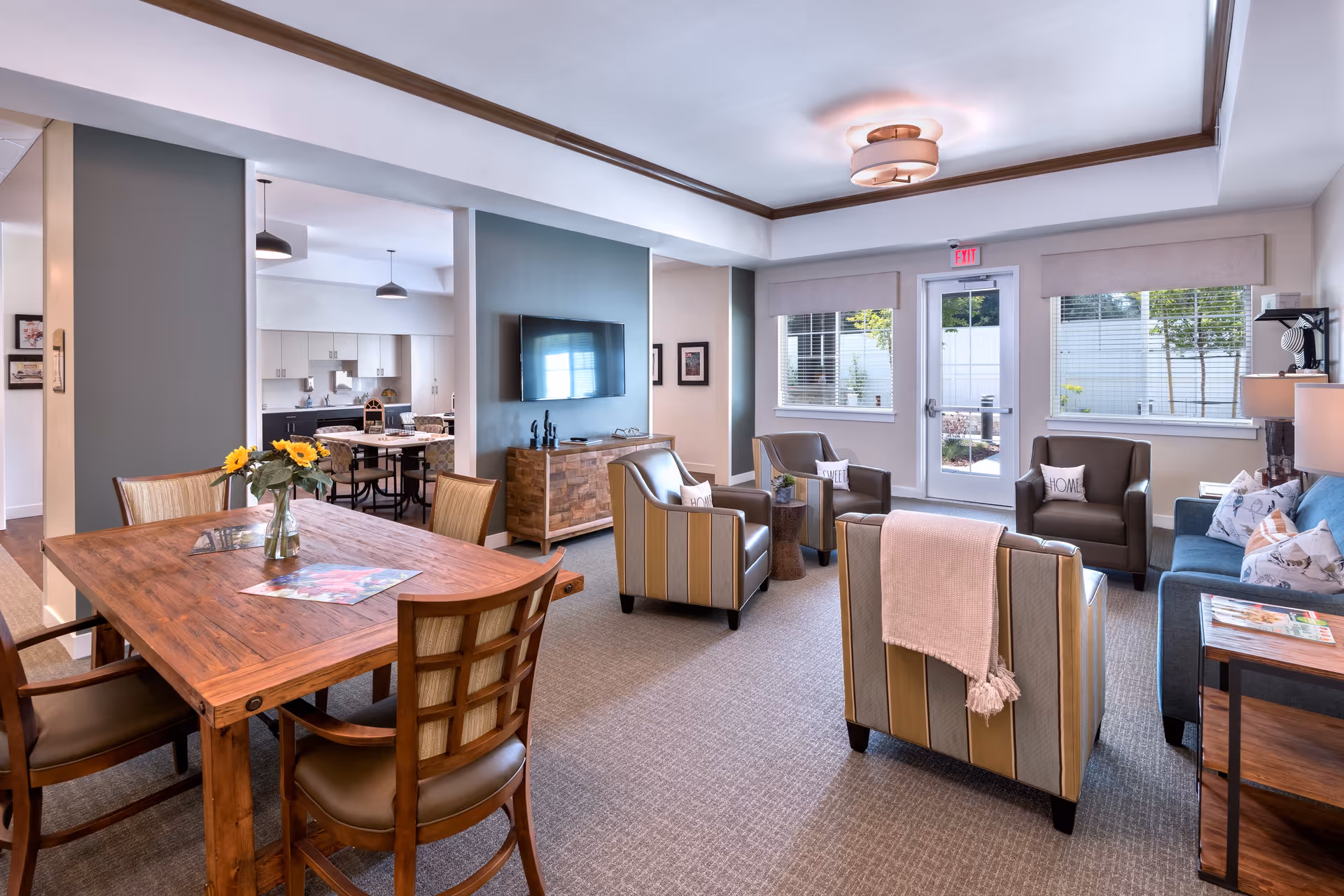 A bright and spacious living room area with a wooden dining table and four chairs on the left, and a seating area on the right featuring a blue sofa, three armchairs with pillows, and a wooden side table. A flat-screen TV is mounted on a wall above a wooden cabinet. Large windows and a glass door provide natural light and a view of an outdoor area. The room has neutral tones with green accent walls and carpeted flooring.