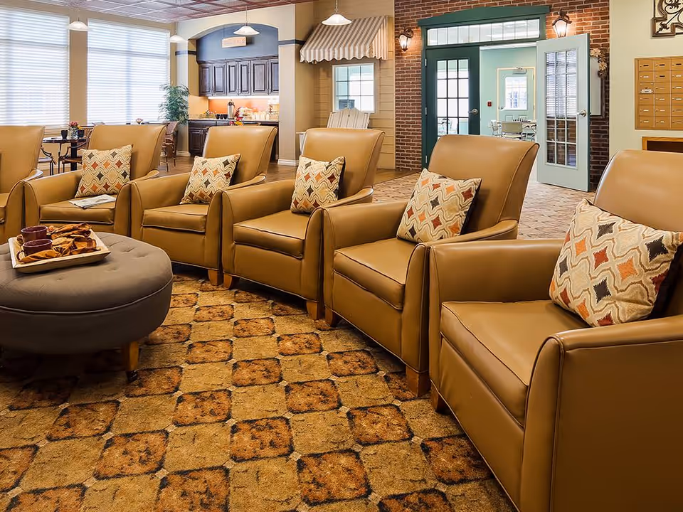 Sunlit communal lounge with a row of tan leather armchairs, patterned pillows, an ottoman with a snack tray, and a kitchenette and entry area in the background.