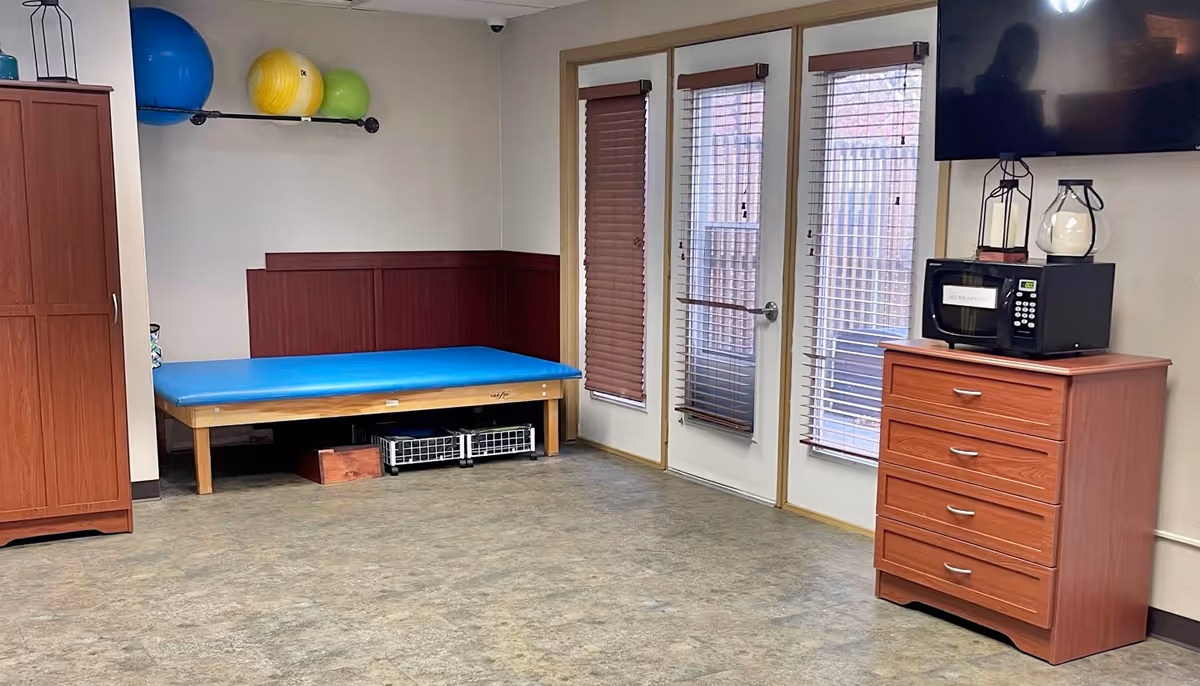 A therapy or exercise room with a blue padded treatment table, three exercise balls on a wall-mounted rack, a wooden cabinet, a chest of drawers with a microwave and decorative items on top, and a wall-mounted TV. There are three tall windows with blinds and a door leading outside.