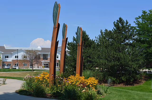 Tall rust-colored metal sculptures rise from a flowerbed of yellow blooms on a landscaped lawn with a multi-story brick-and-white building and trees in the background.