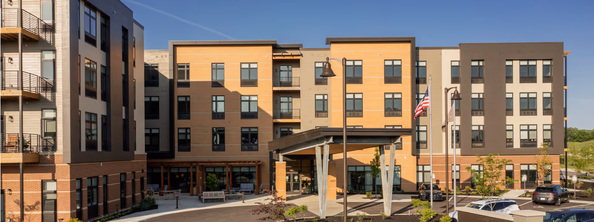 Front entrance of a multi-story senior living building with a covered porte-cochere, American flag, and parked cars.