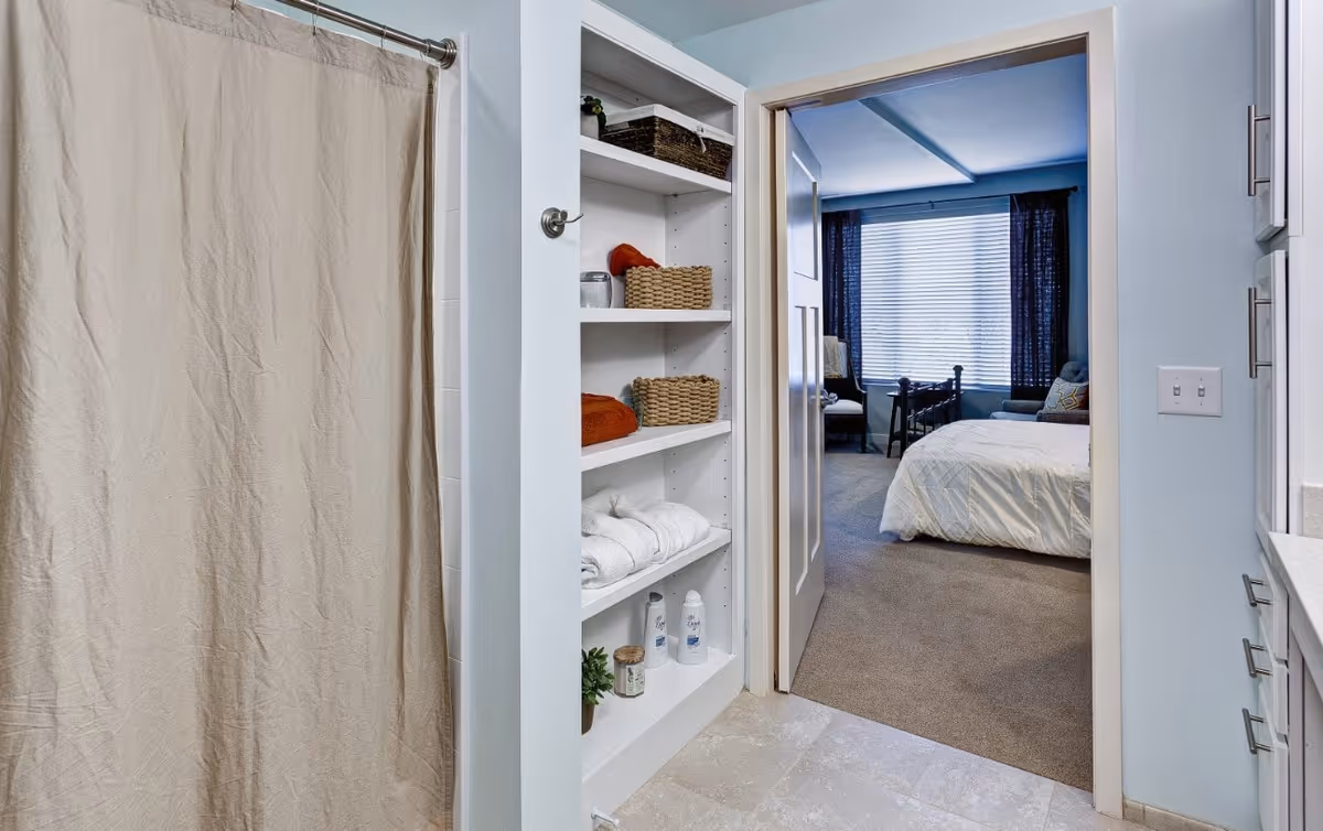 View from a bathroom showing a shower with a beige curtain, built-in shelves with towels and baskets, and a doorway leading into a bedroom.