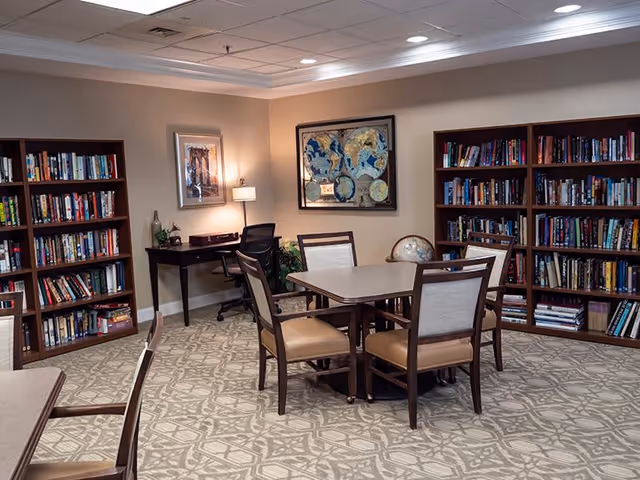 A cozy library room with two large wooden bookshelves filled with books, a square table surrounded by four chairs with cushioned seats, a desk with a black office chair, a lamp, and framed artwork on the beige walls.