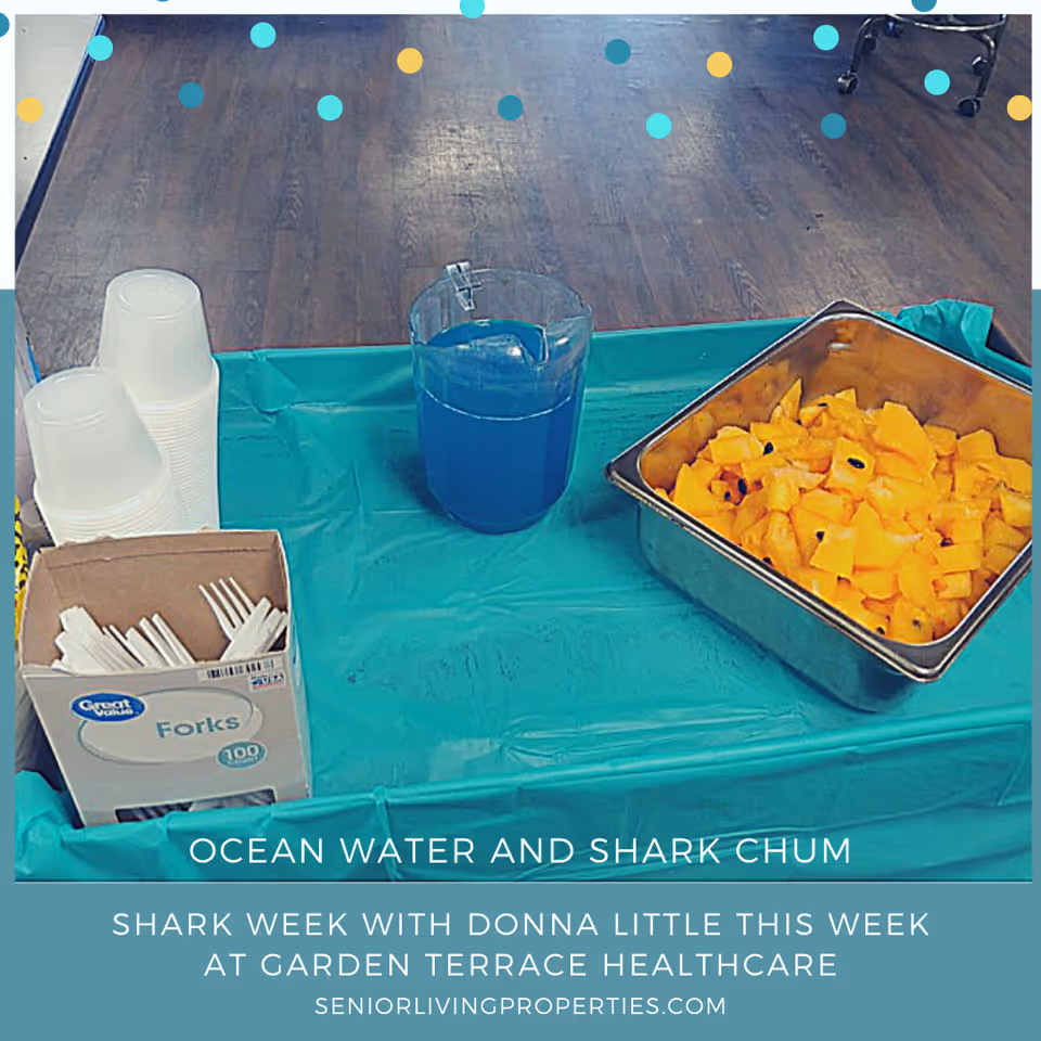Table with a pitcher of blue punch, a tray of cubed fruit, stacked plastic cups and a box of forks on a teal tablecloth with Shark Week promotional text at the bottom.