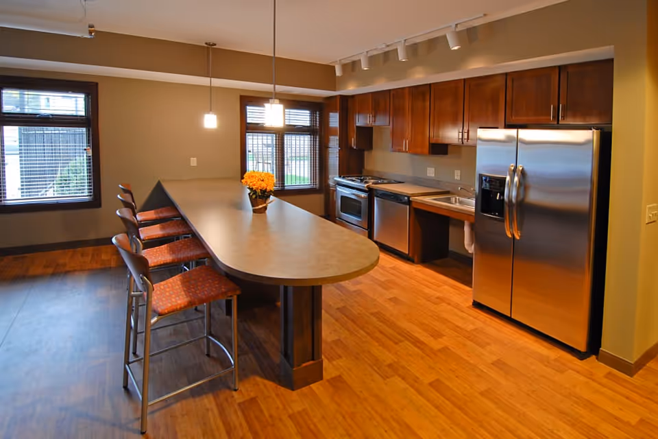Modern kitchen area with wooden cabinets, stainless steel refrigerator, stove, dishwasher, and a long island countertop with four high chairs. There are two pendant lights hanging above the island and windows with blinds letting in natural light.