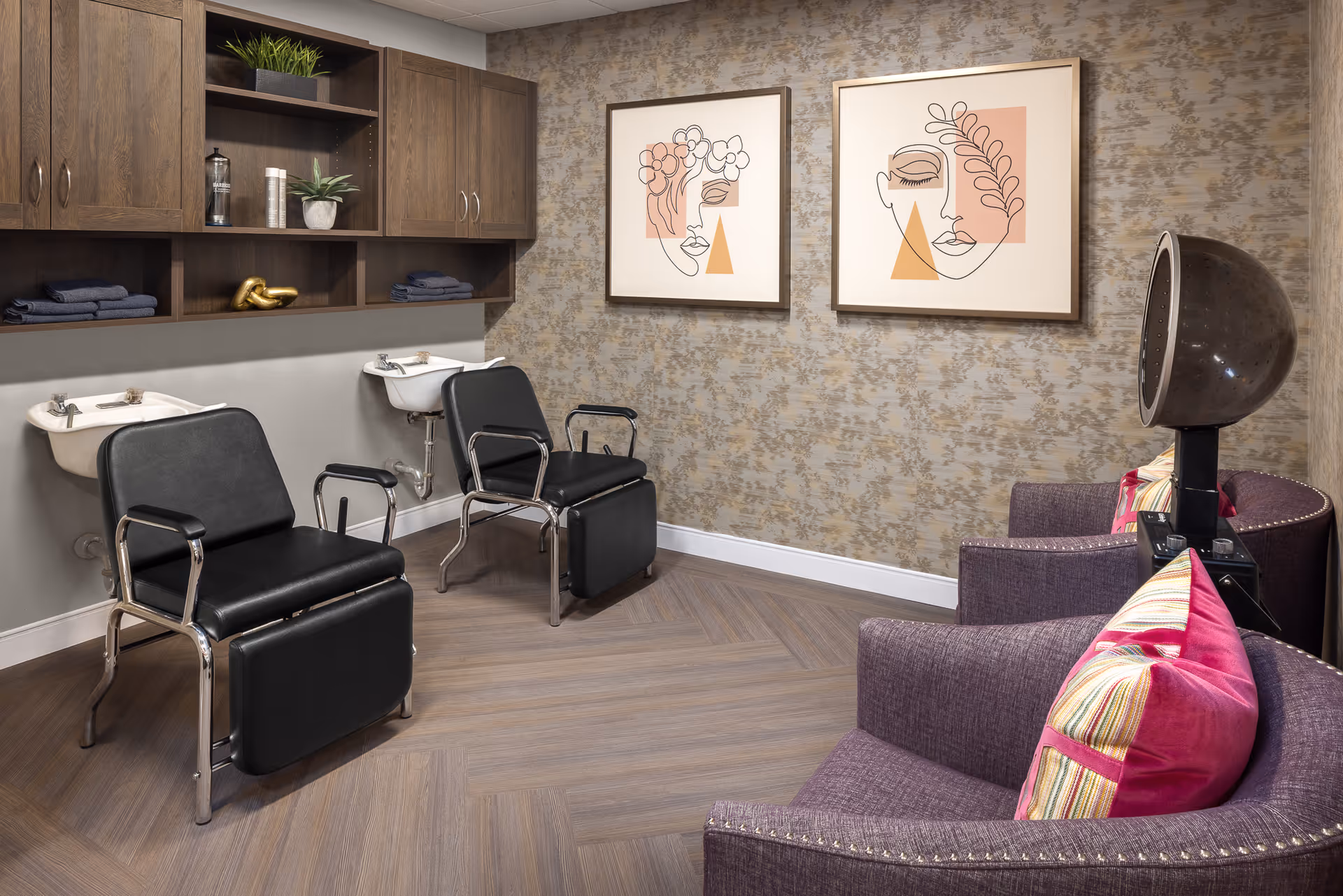 Interior of a senior living facility hair salon area with two black salon chairs in front of two white sinks. There are wooden cabinets with plants and towels above the sinks. On the right side, there are two purple armchairs with colorful pillows and a vintage hair dryer. The wall has patterned wallpaper and two framed abstract face artworks.