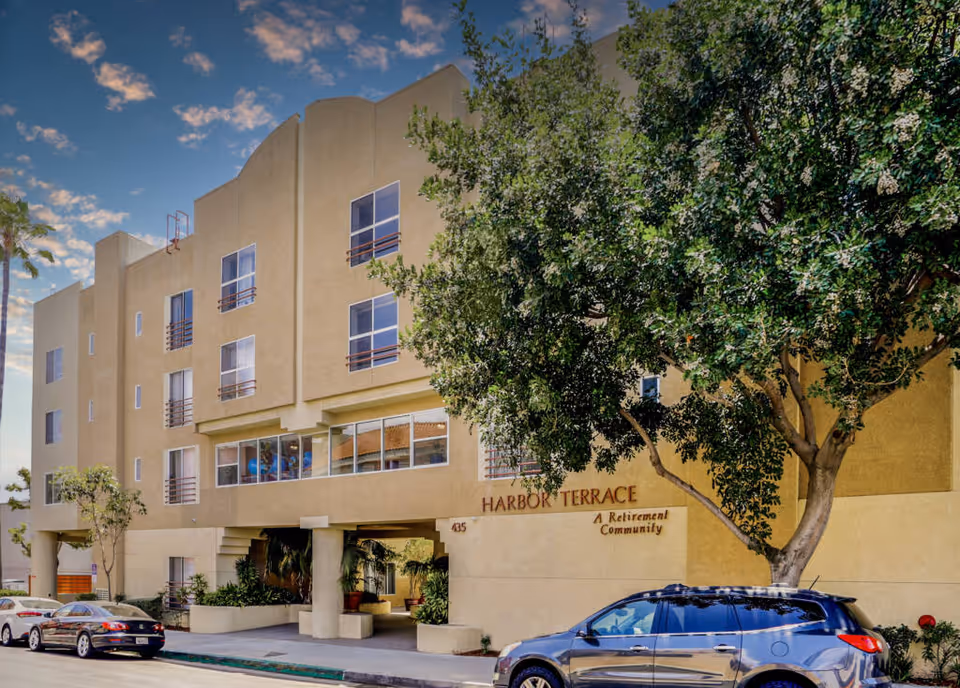 Exterior view of Harbor Terrace Retirement Community building with several windows, a large tree in front, and parked cars along the street under a partly cloudy sky.