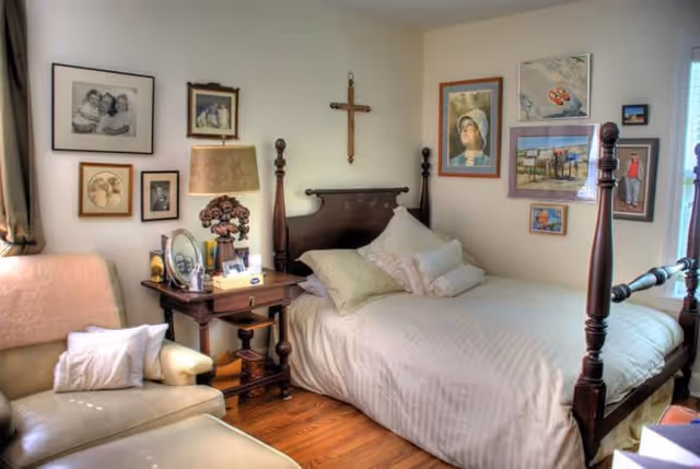 A cozy bedroom with a wooden four-poster bed made with white linens and multiple pillows. Next to the bed is a wooden nightstand with a lamp, framed photos, and decorative items. A comfortable armchair with a pillow is positioned near the bed. The walls are decorated with various framed pictures and artwork, and a wooden cross hangs above the bed. The room has wooden flooring and a window letting in natural light.