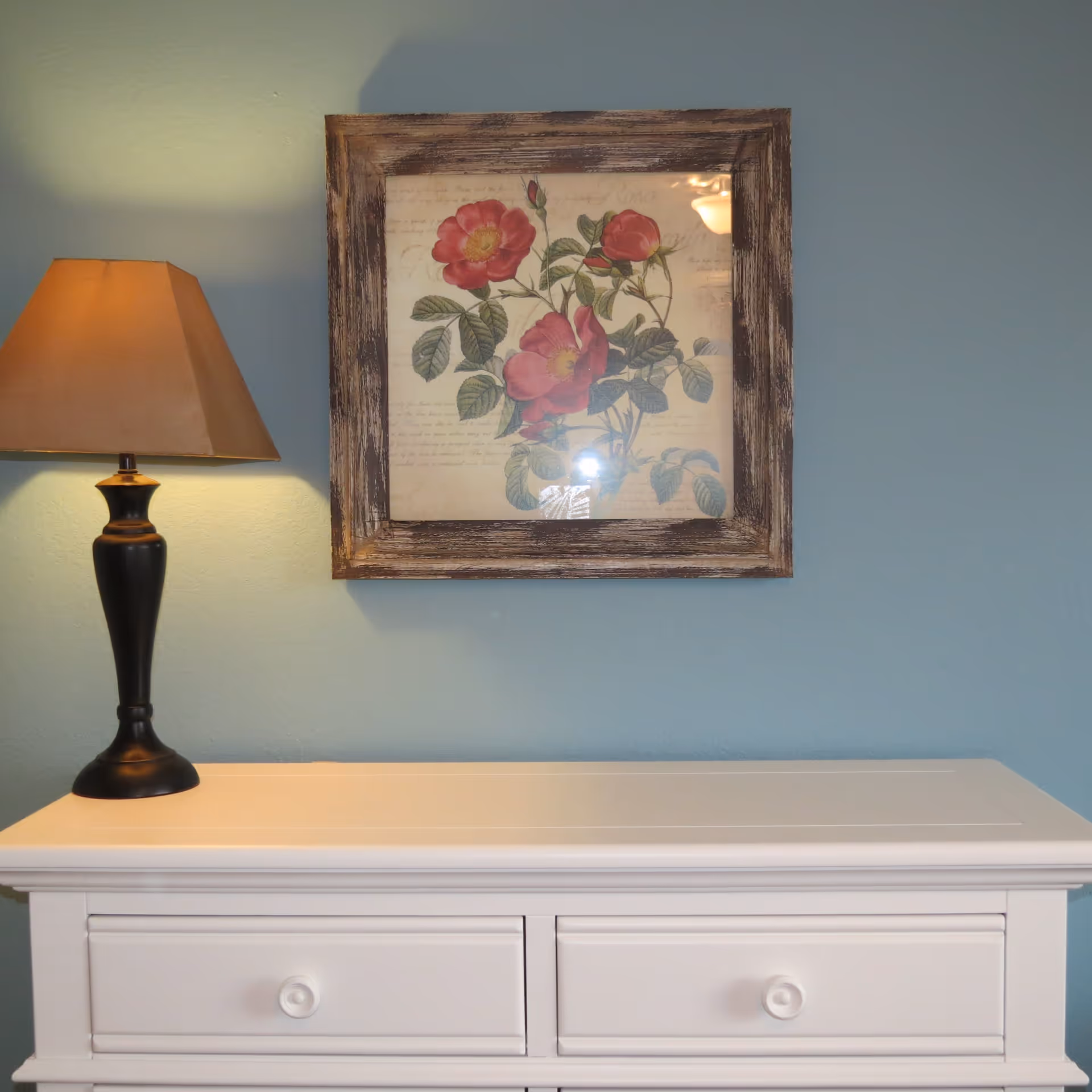 White dresser topped by a lamp beneath a framed floral print on a blue wall.
