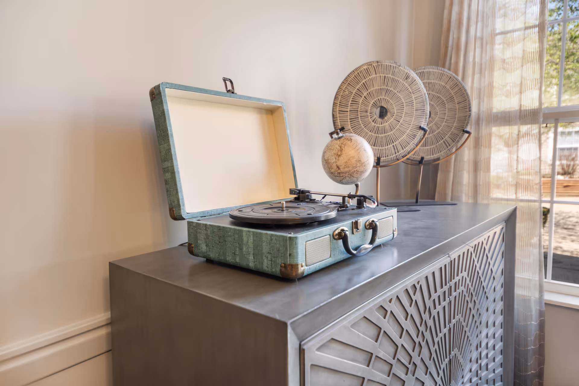 A vintage-style portable record player with a greenish-blue case sits open on a decorative metal cabinet. Behind it are two round woven decorative plates on stands and a small globe. A window with sheer curtains allows natural light into the room.