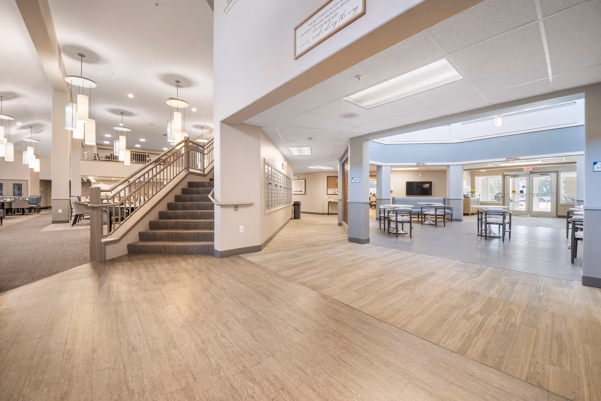 Interior view of a senior living facility showing a spacious common area with a staircase leading to an upper floor, multiple hanging pendant lights, seating areas with tables and chairs, a hallway with mailboxes on the wall, and large windows letting in natural light.
