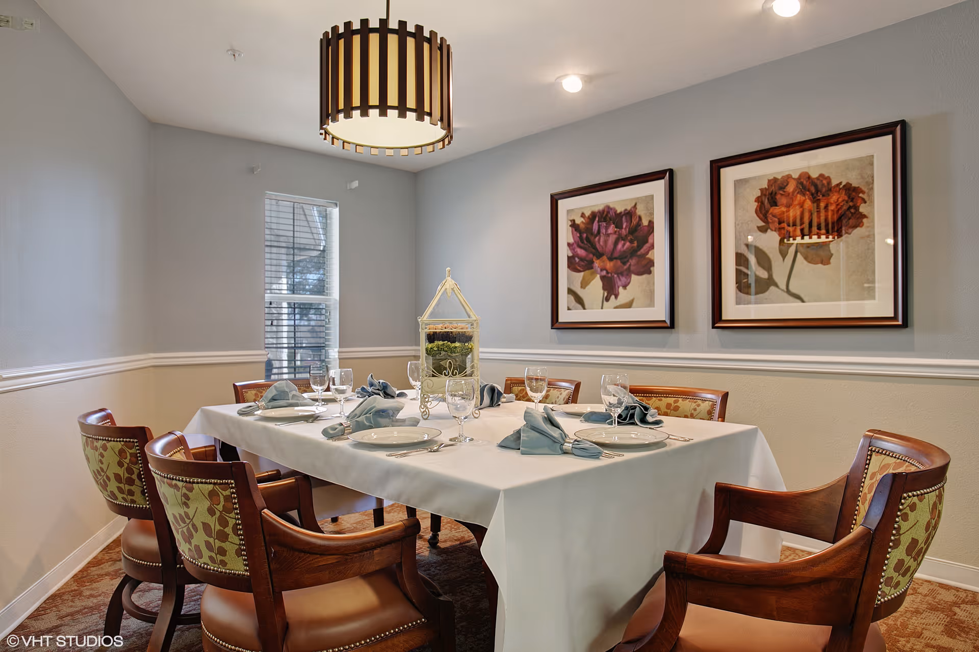 A dining room with a rectangular table covered with a white tablecloth, set with plates, glasses, silverware, and folded blue napkins. Six wooden chairs with patterned upholstery surround the table. Two framed floral paintings hang on the wall, and a modern pendant light fixture hangs from the ceiling. A window with blinds is visible in the background.