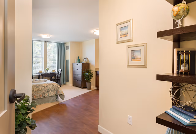 View into a well-lit studio apartment showing a bed, dresser, small dining table by large windows, and decorative shelving in the foreground.