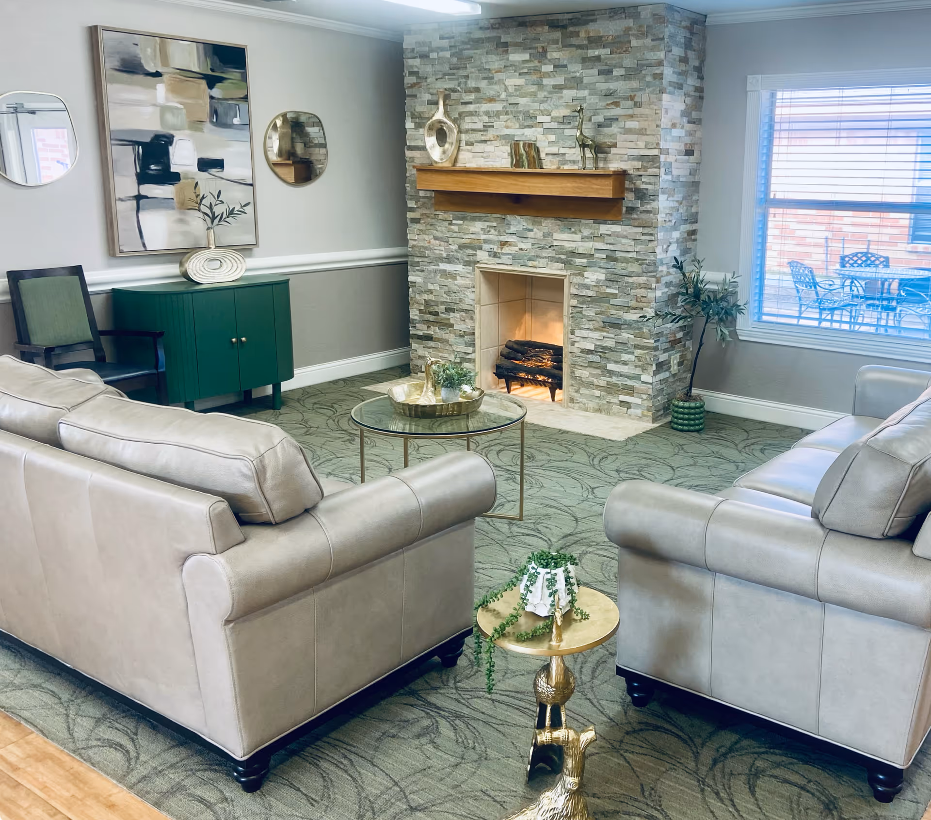 A cozy living room with two beige leather sofas facing a stone fireplace with a wooden mantle. The room has a green patterned carpet, a round glass coffee table with decorative items, and a small gold side table with a plant. On the wall above a green cabinet are two round mirrors and an abstract painting. A large window with blinds shows an outdoor patio with a table and chairs.