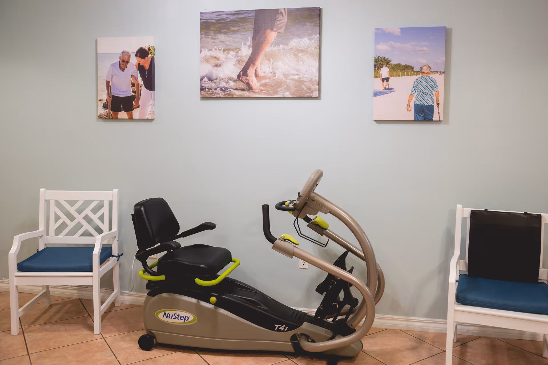 A small exercise area with a NuStep T41 recumbent cross trainer positioned between two white chairs with blue cushions. Three framed photos of elderly people enjoying outdoor activities are hung on the light green wall above the exercise equipment.