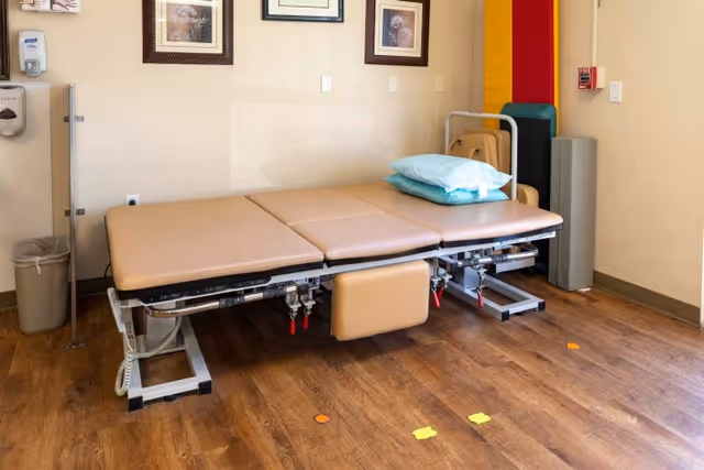 A therapy or examination room with a beige adjustable treatment table with two blue pillows on it. The room has wooden flooring, a trash can, a hand sanitizer dispenser, and framed pictures on the wall. There are also folded chairs and exercise mats stored in the corner.
