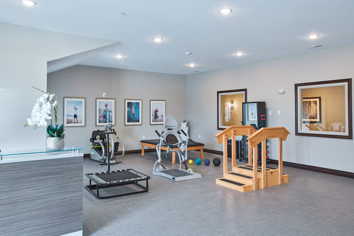 A well-lit exercise room in a senior living facility featuring various fitness equipment including a recumbent exercise bike, a small trampoline, therapy balls, and wooden parallel bars. The room has light gray walls adorned with framed pictures and two large windows with dark brown frames. A reception desk with a white orchid plant in a silver pot is visible on the left side.