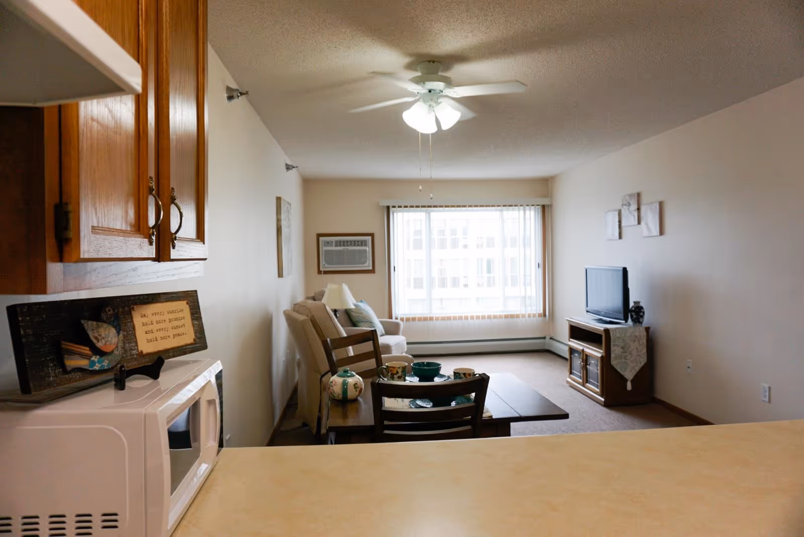 View from a kitchen counter into a living room area with a ceiling fan, a window with vertical blinds, a beige couch, a wooden dining table with chairs, a teapot and cups, and a TV on a wooden stand with decorative items.