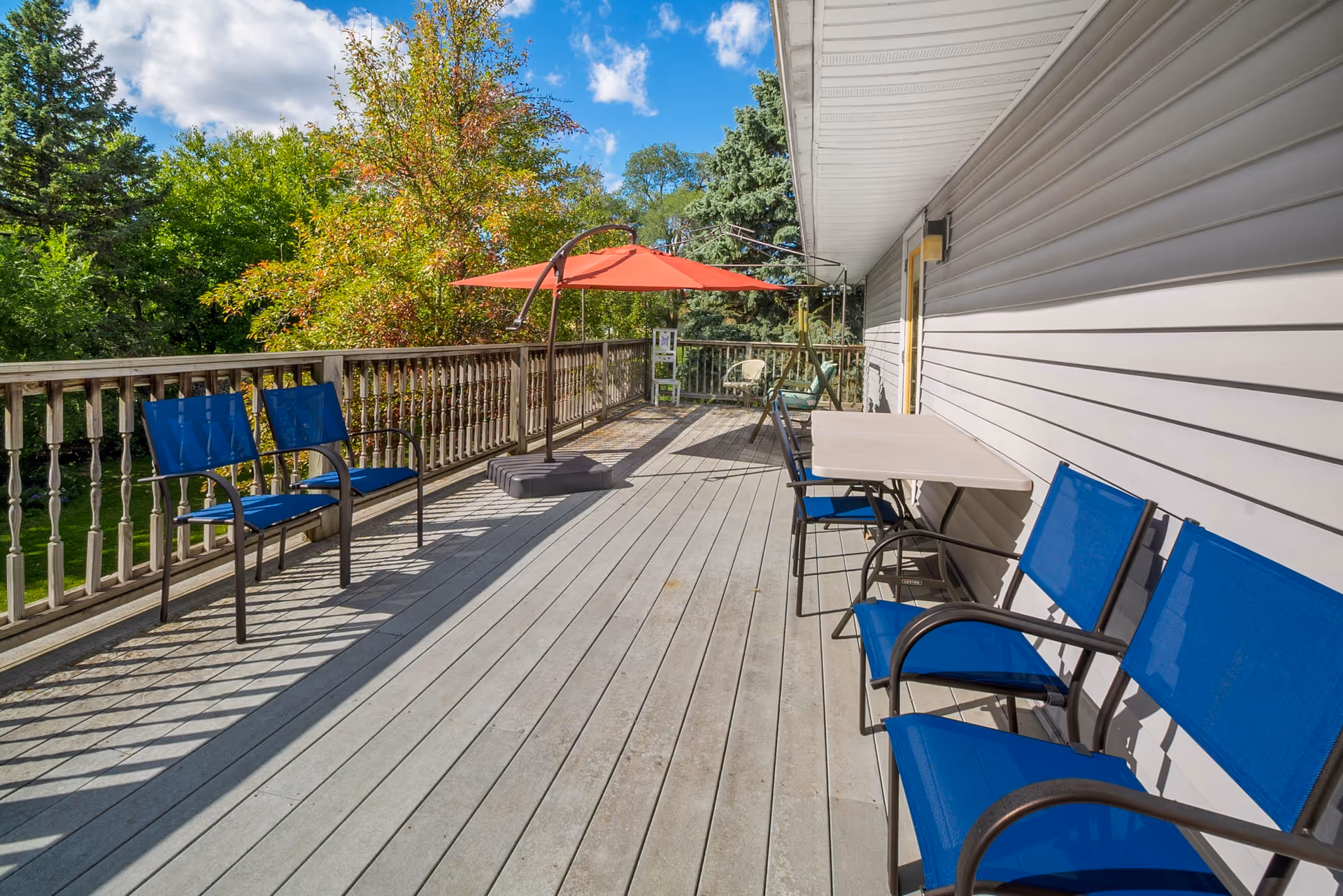 Outdoor wooden deck attached to a building with blue chairs and a table along the wall. There is a red umbrella providing shade and green trees surrounding the deck under a partly cloudy sky.