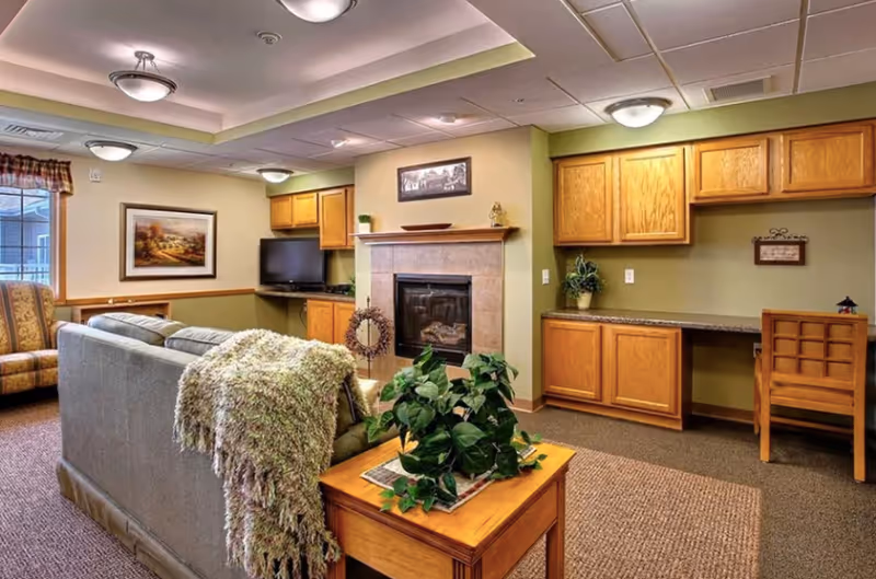 A cozy senior living room featuring a gray sofa with a textured throw blanket, a wooden coffee table with a green leafy plant, a fireplace with a mantel, a flat-screen TV mounted on the wall, wooden cabinets, a small desk with a chair, and a window with a valance letting in natural light.