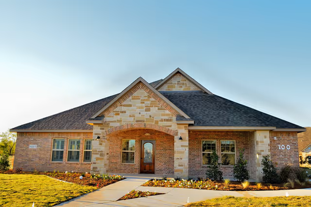 Front exterior view of a single-story brick and stone building with a peaked roof, a wooden front door with glass panels, multiple windows, and a landscaped lawn with a concrete walkway leading to the entrance.