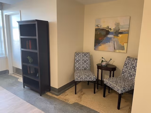 A small seating area with two patterned chairs and a round wooden side table between them, holding a decorative item. Behind the chairs is a wall with an abstract painting. To the left, there is a tall dark wooden bookshelf with some books and a small plant. The floor has a mix of carpet and tile, and there is a door with a window on the far left.