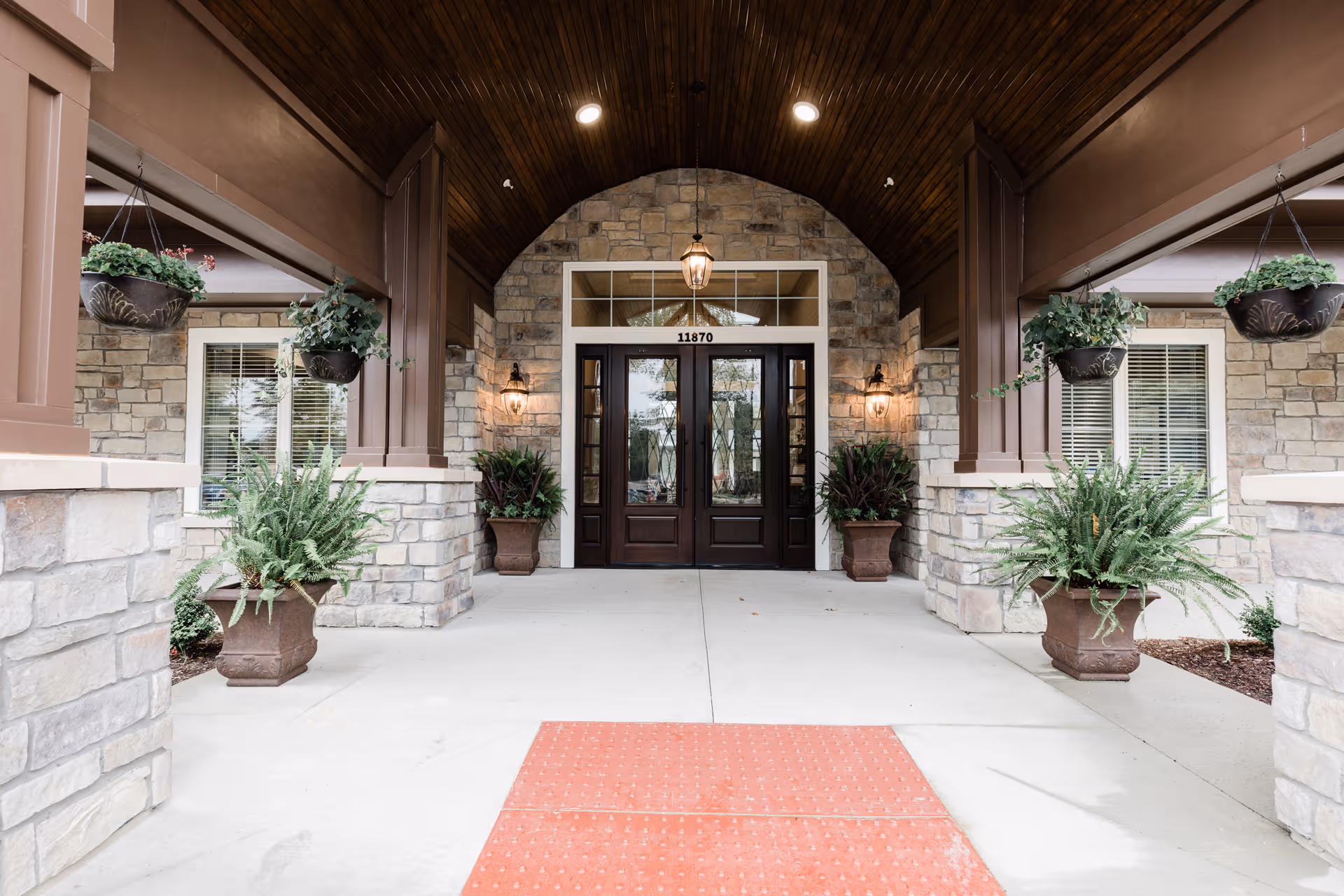 Entrance of a building with stone walls and a wooden arched ceiling. There are double dark wooden doors with glass panels, flanked by two wall-mounted lantern-style lights. The entrance is decorated with potted plants and hanging flower baskets. The address number 11870 is displayed above the doors.