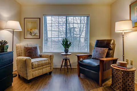 A cozy sitting area with two armchairs, one beige patterned and one black leather with wooden armrests, positioned on either side of a small round table holding a potted plant. The room has wooden flooring, a large window with blinds, two floor lamps providing warm light, and framed artwork on the walls.
